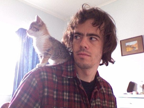 A photo of a young man with terrible hair and a great flannel shirt and a small calico kitten perched on his shoulder.